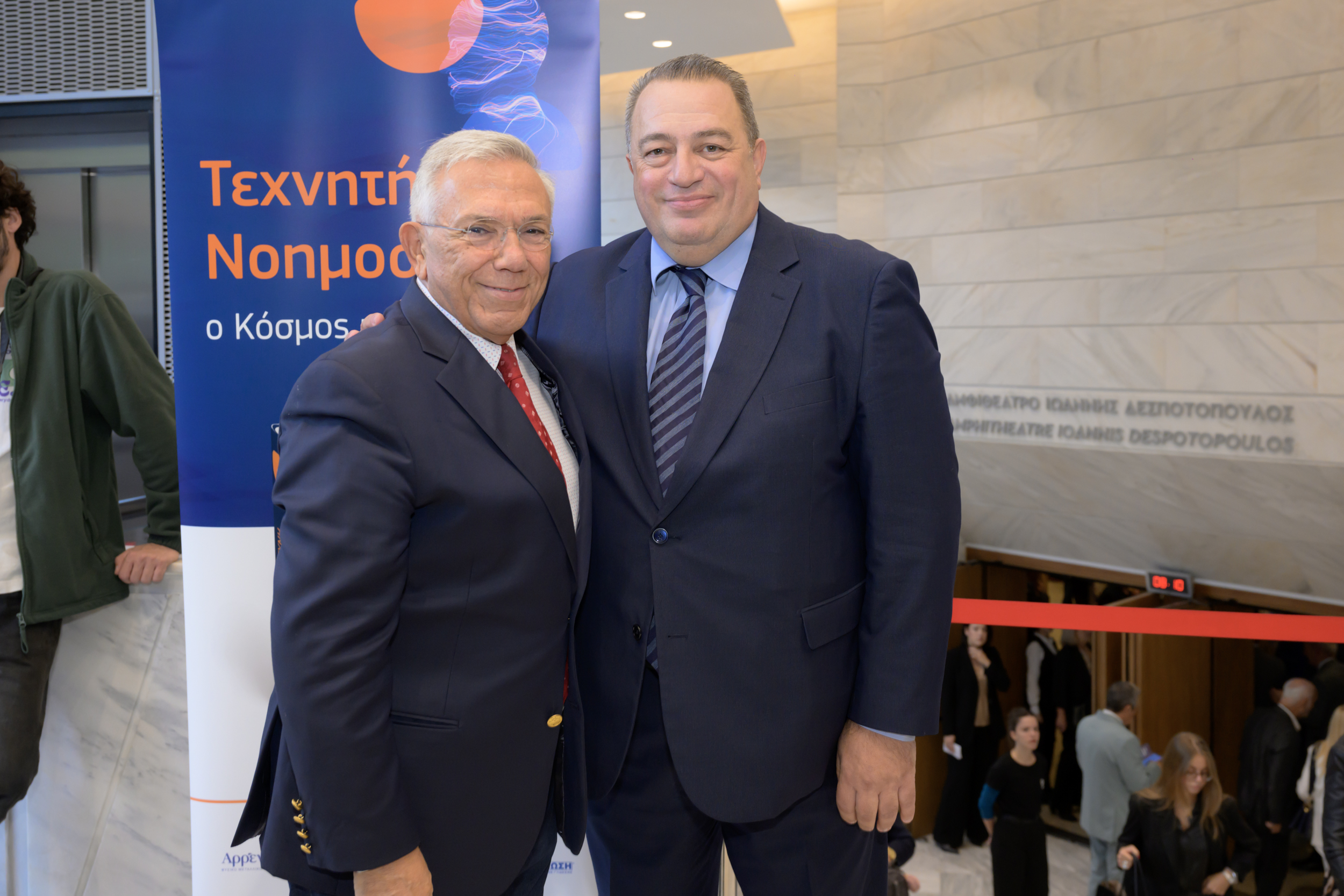 Δύο άτομα φωτογραφίζονται δίπλα σε πανό στον χώρο υποδοχής.