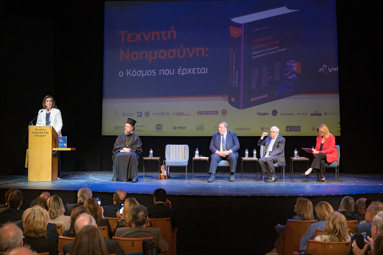 Ομιλήτρια στο αναλόγιο, καθιστοί συμμετέχοντες στη σκηνή και κοινό στο προσκήνιο.