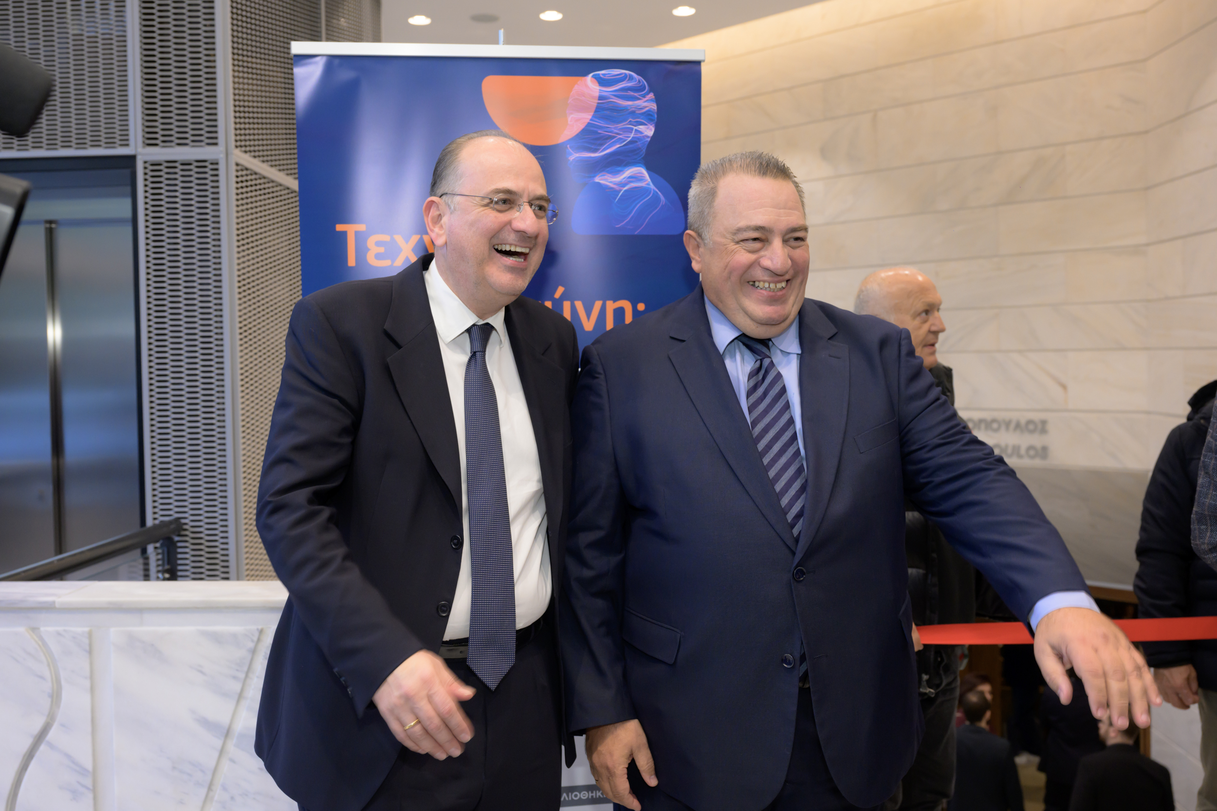 Δύο άτομα χαμογελούν στον χώρο υποδοχής με το πανό της εκδήλωσης πίσω τους.