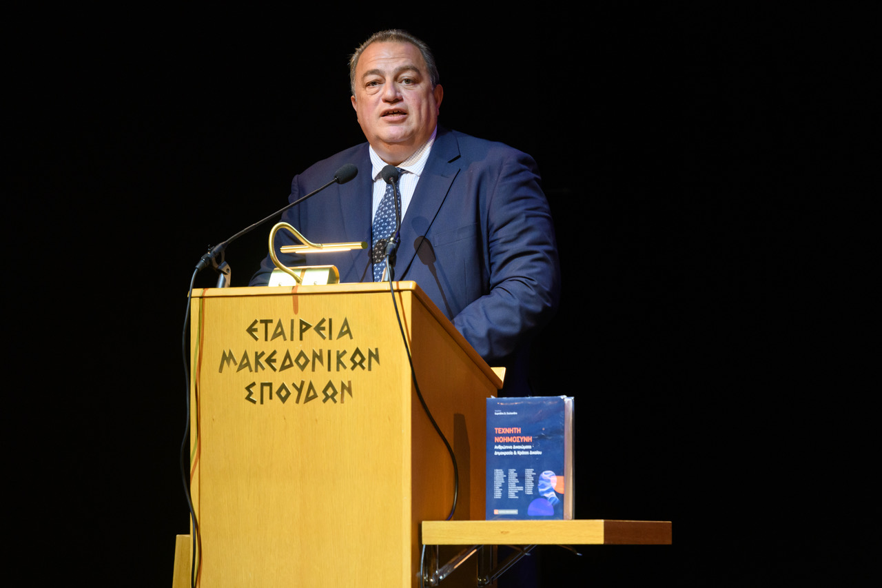 Ομιλητής σε ξύλινο αναλόγιο με μικρόφωνα και βιβλίο δίπλα του.